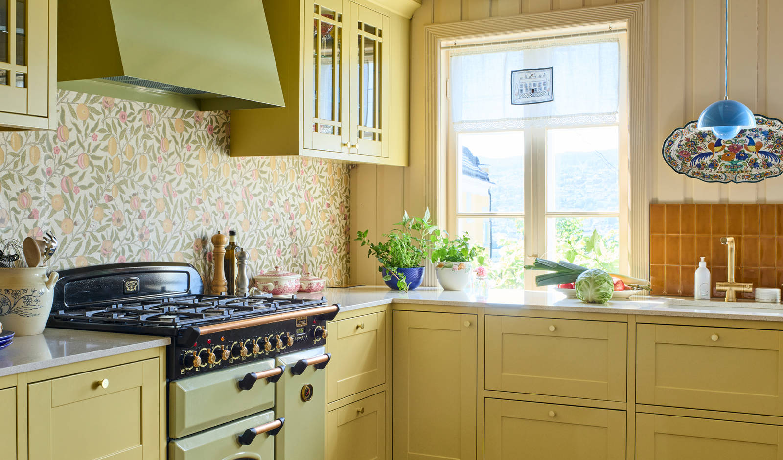 Landlig rammekjøkken Scala Inframe fra Sigdal som gir en snekkerbygged preg, vitrineskaber, Falcon Classic Olive green komfyr med gassbrennere og Røroshetta Grue ventilator i samma fargekode, messing knotter og blandebatteri