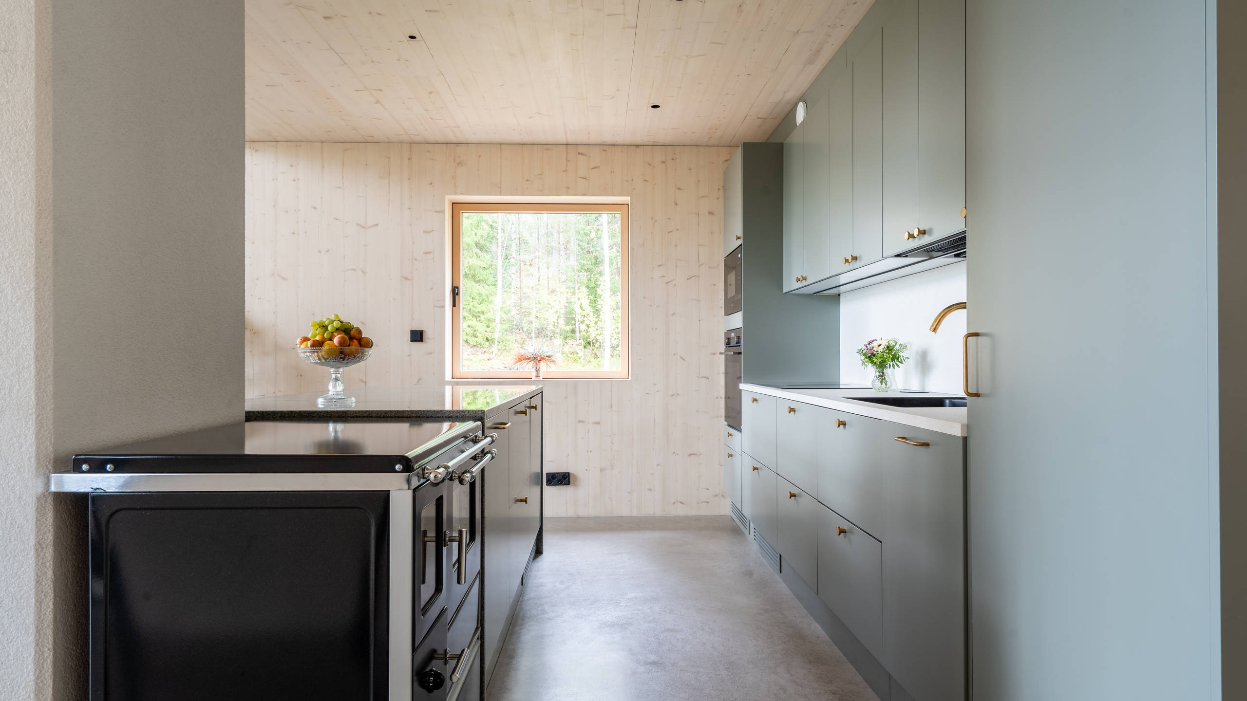 Vihreä keittiö Novartin luonnonvärisessä Havu-sävyssä, messinkinuppeilla, graniittitasoilla ja lasiovilla, joissa on alumiinikehykset keittiön altaassa.