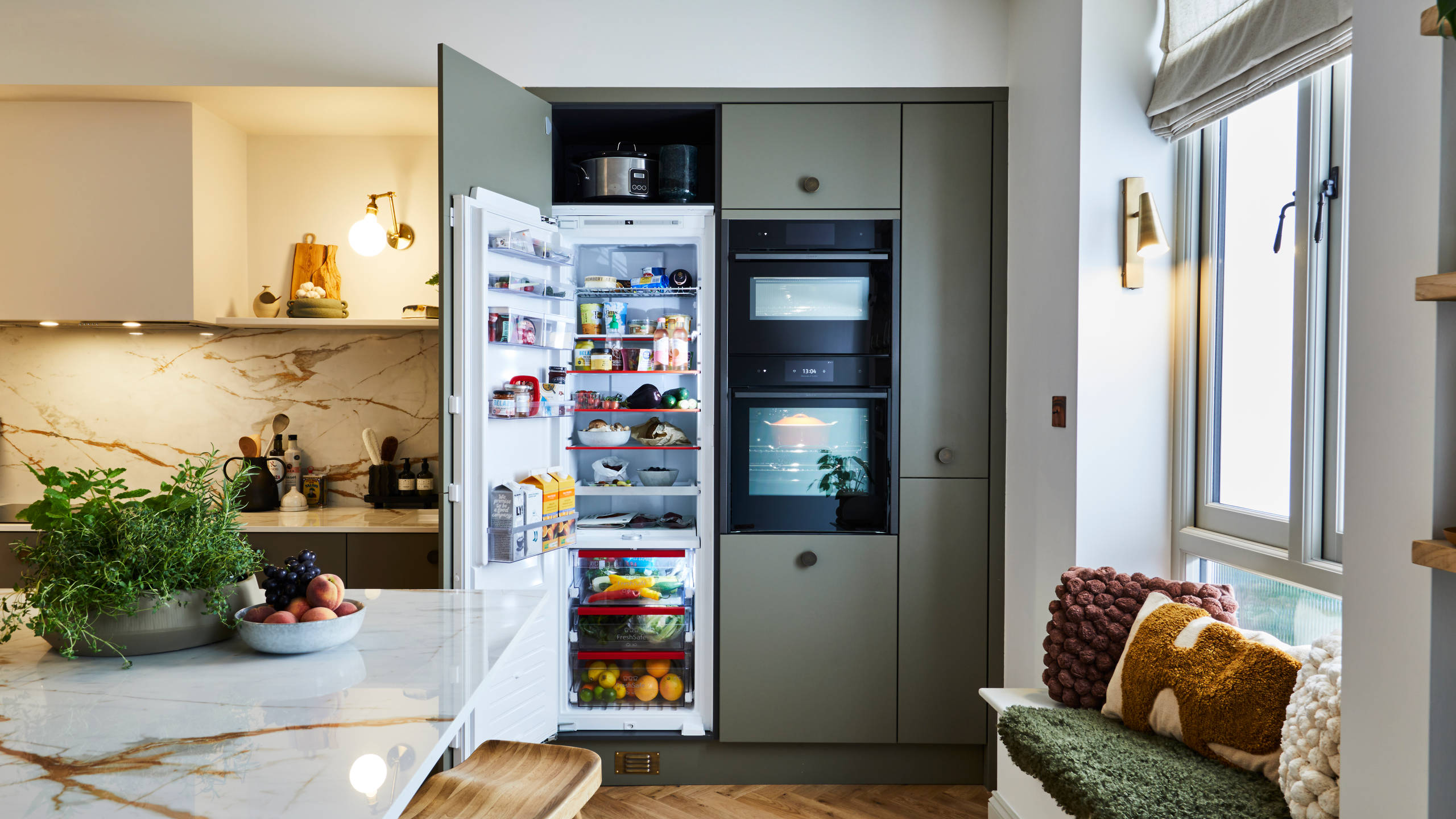 Open fridge in Soho Balmoral tall cabinets with integrated ovens and a bench in the window, next to a wall of open wood shelves decorated with plants, candles and wine glasses.