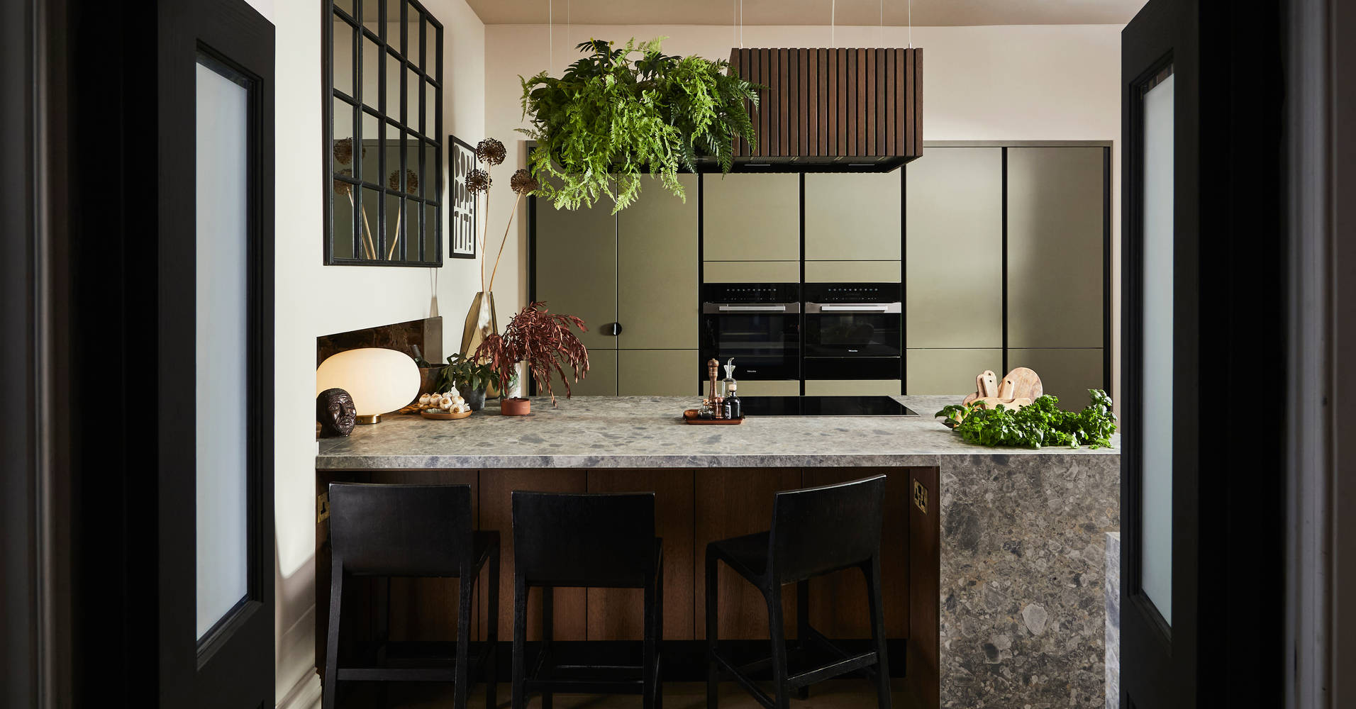 View through opened dark wood doors of the biophilic Dramatic neutral Balmoral Soho and Nordic Nature kitchen from Magnet and matching dark chairs by the bar area of the kitchen island.