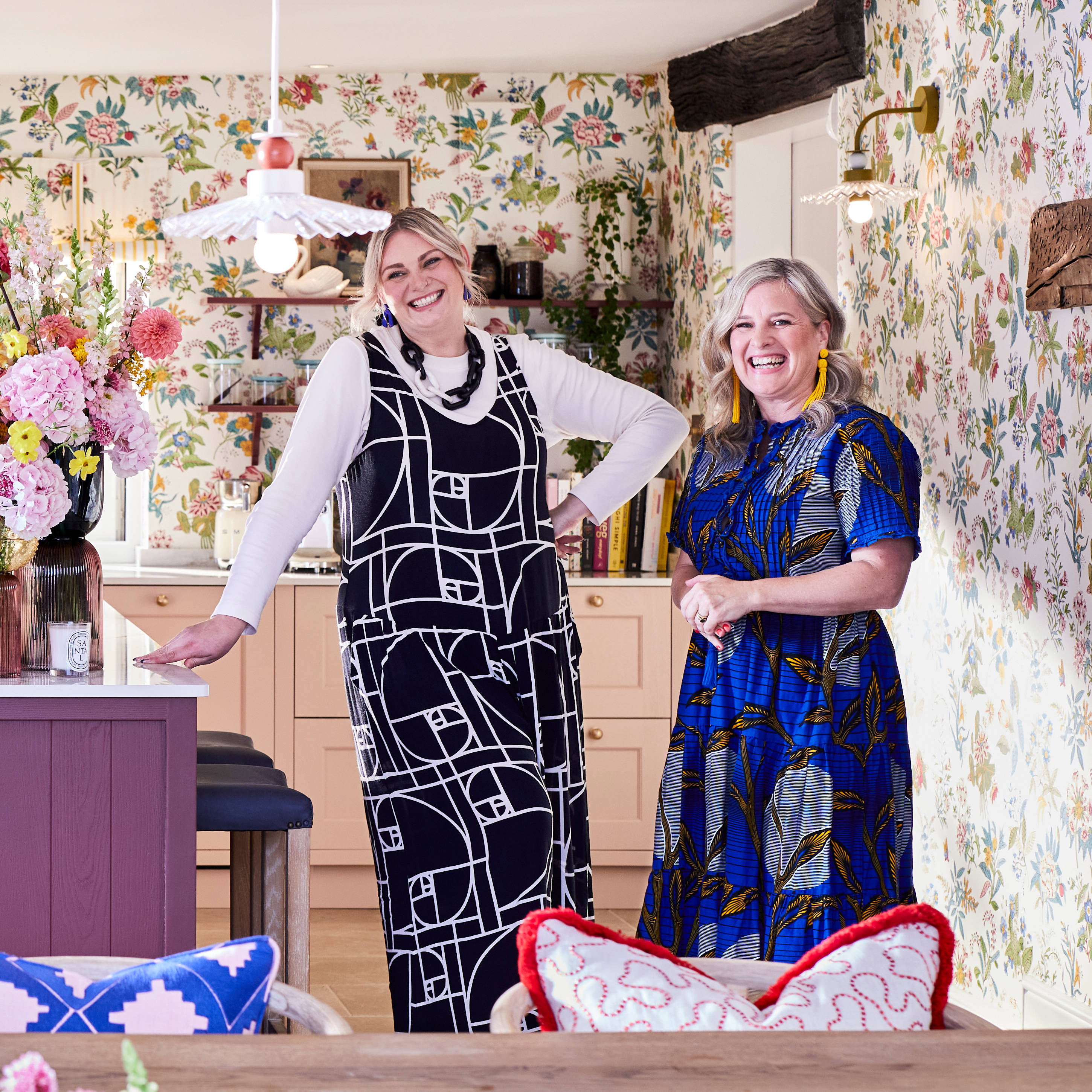 Full room view of Jen from Magnet and Sophie Robinson laughing standing next to the kitchen island in the traditional Shaker style Ludlow kitchen in colours Burlington red, Harvest and Chalk blush matched with a maximalist floral wallpaper.
