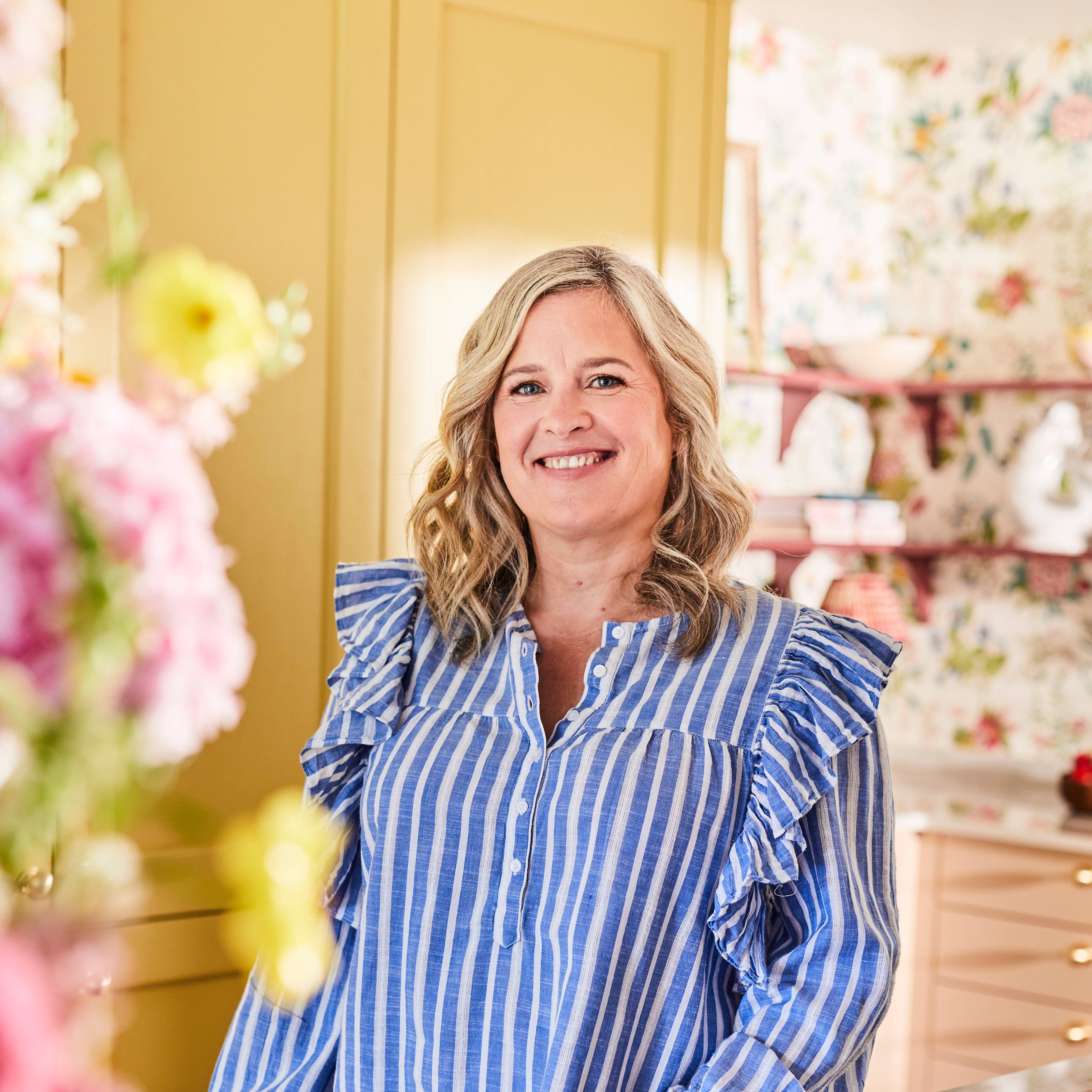Sophie wearing a striped blouse, posing by the kitchen island in her colourful kitchen with Magnet colours harvest and Clalk blush in the background