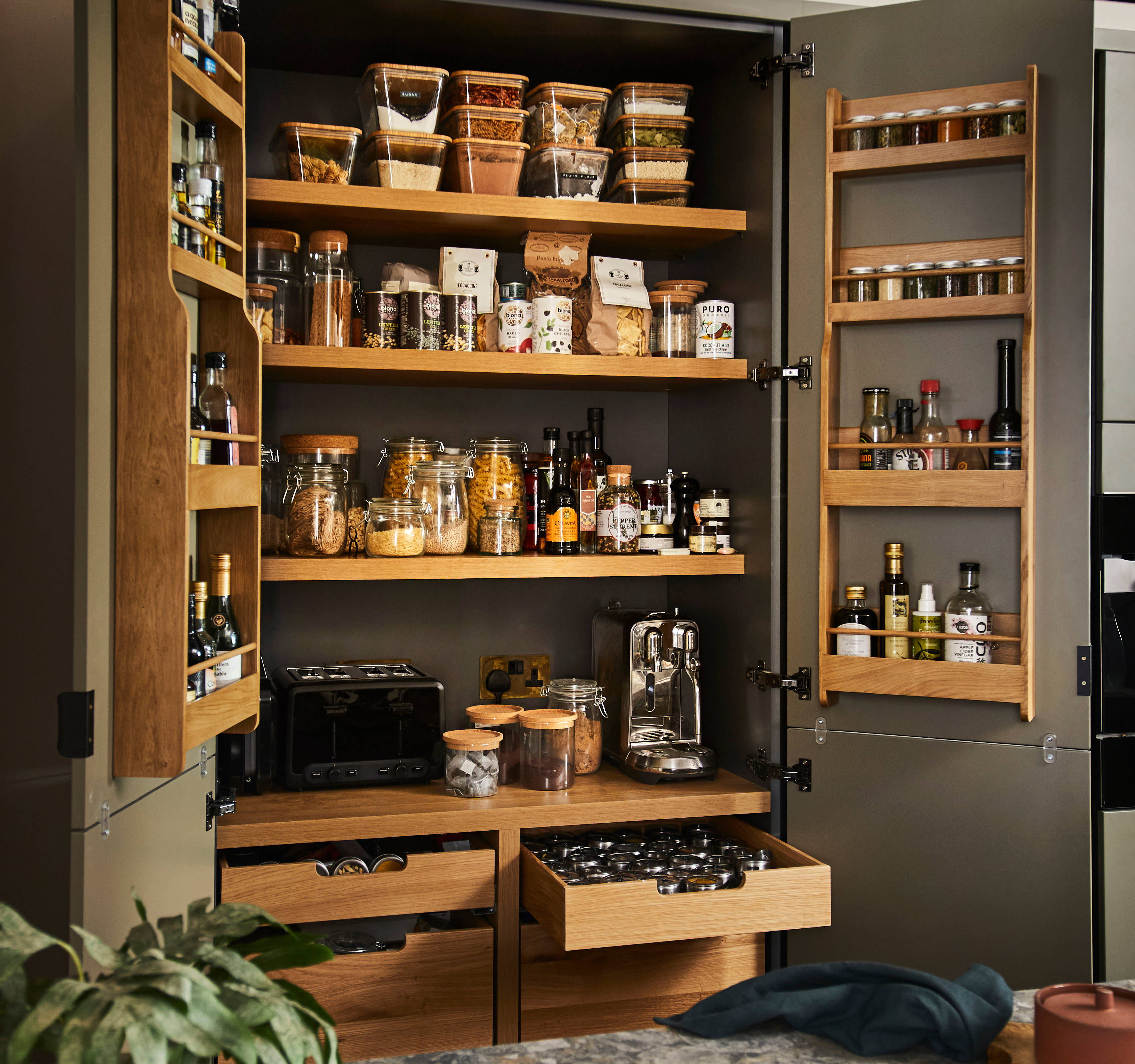 Diagonal view from kitchen island of opened pantry larder with oak interior full of glass jars containing dry goods, a toaster and spices