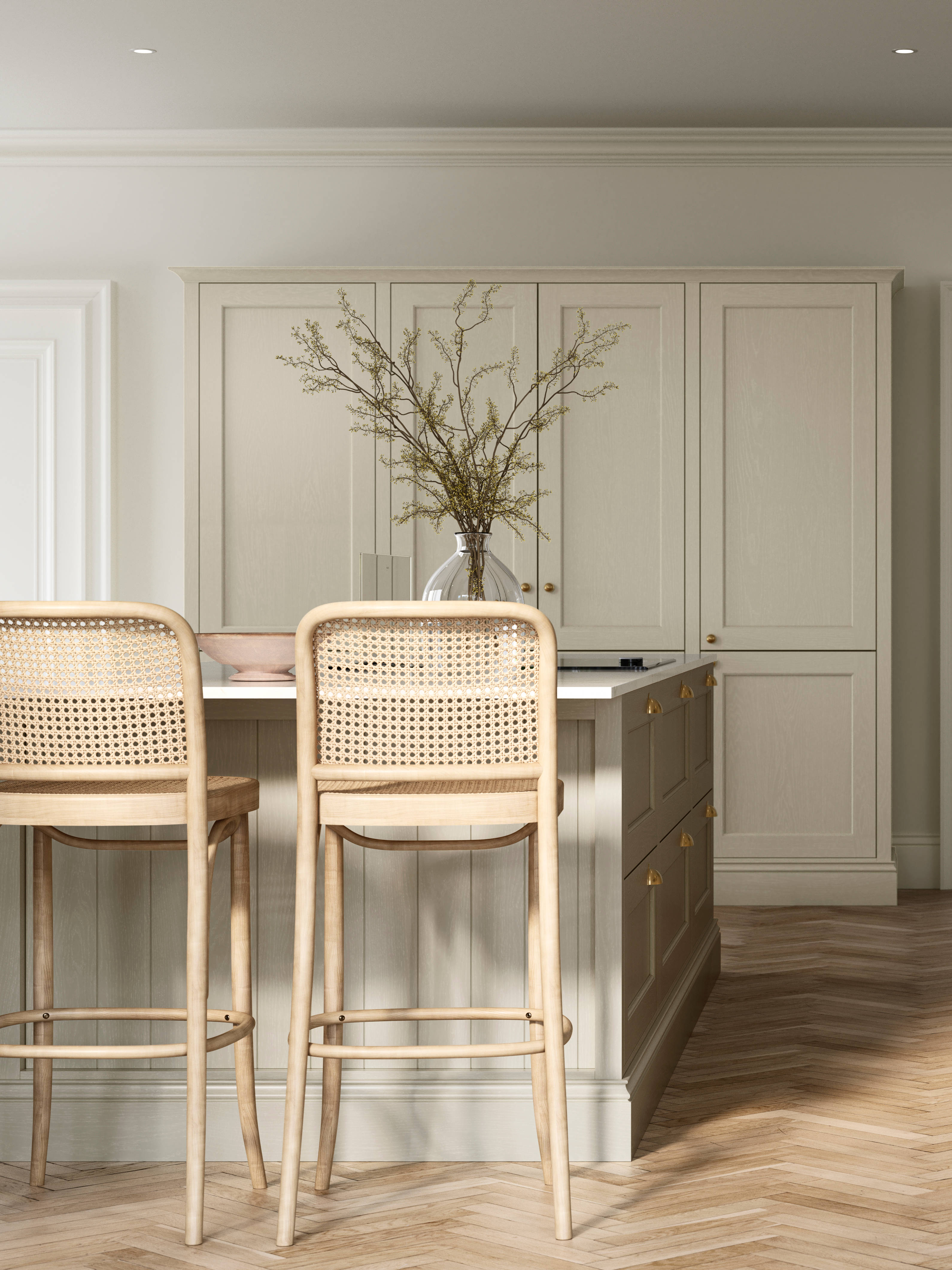 Traditional Shaker-style kitchen Ludlow Parchment with tactile woodgrain, tall cabinets, brass cup handles and knobs, integrated ovens, a kitchen island with rattan bar stools, a vase holding foliage, and light wood herringbone flooring.