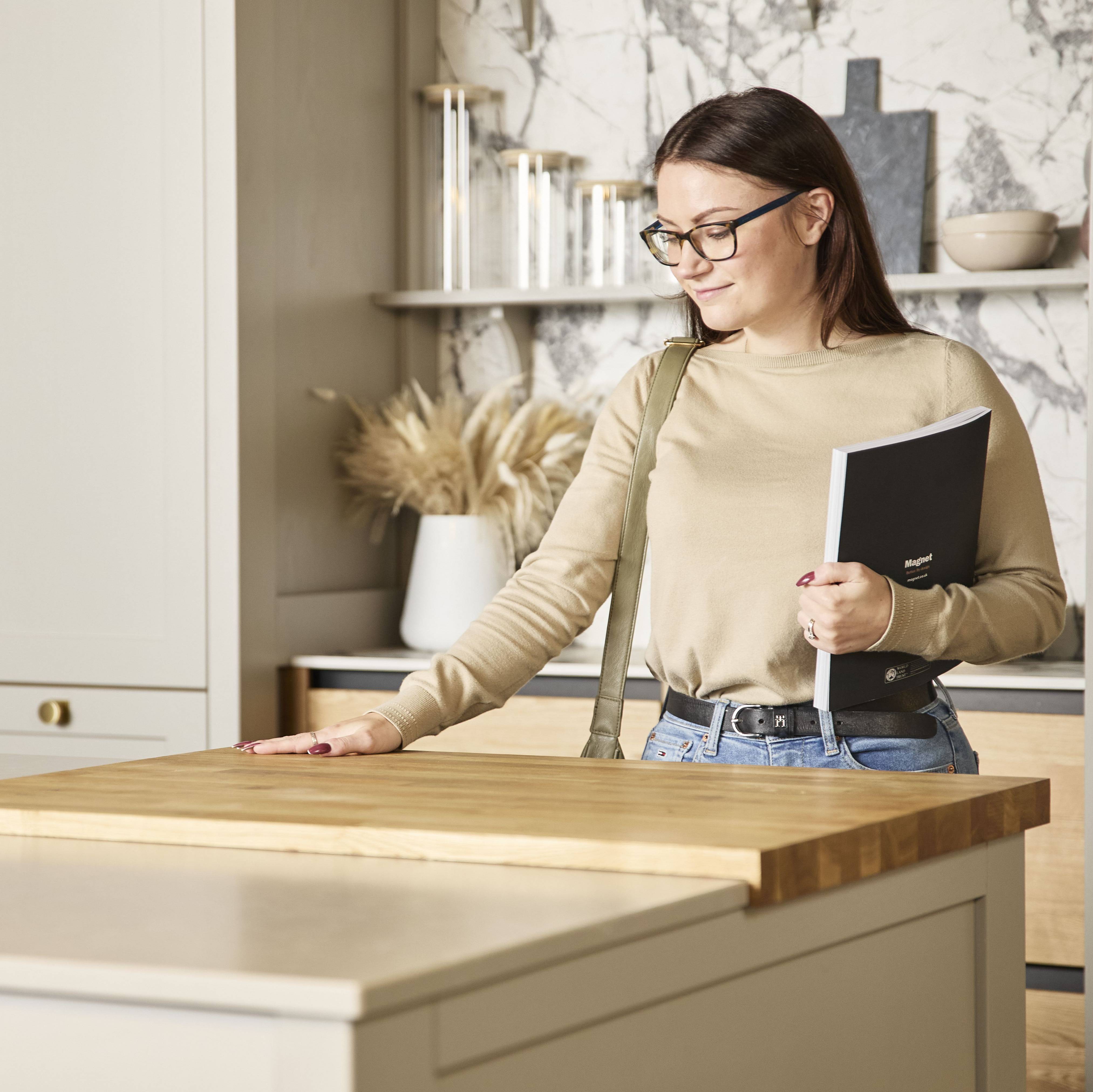 Woman holding a brochure feeling the wood worktop with her hand in a neutral shade Shaker style kitchen in a Magnet Showroom