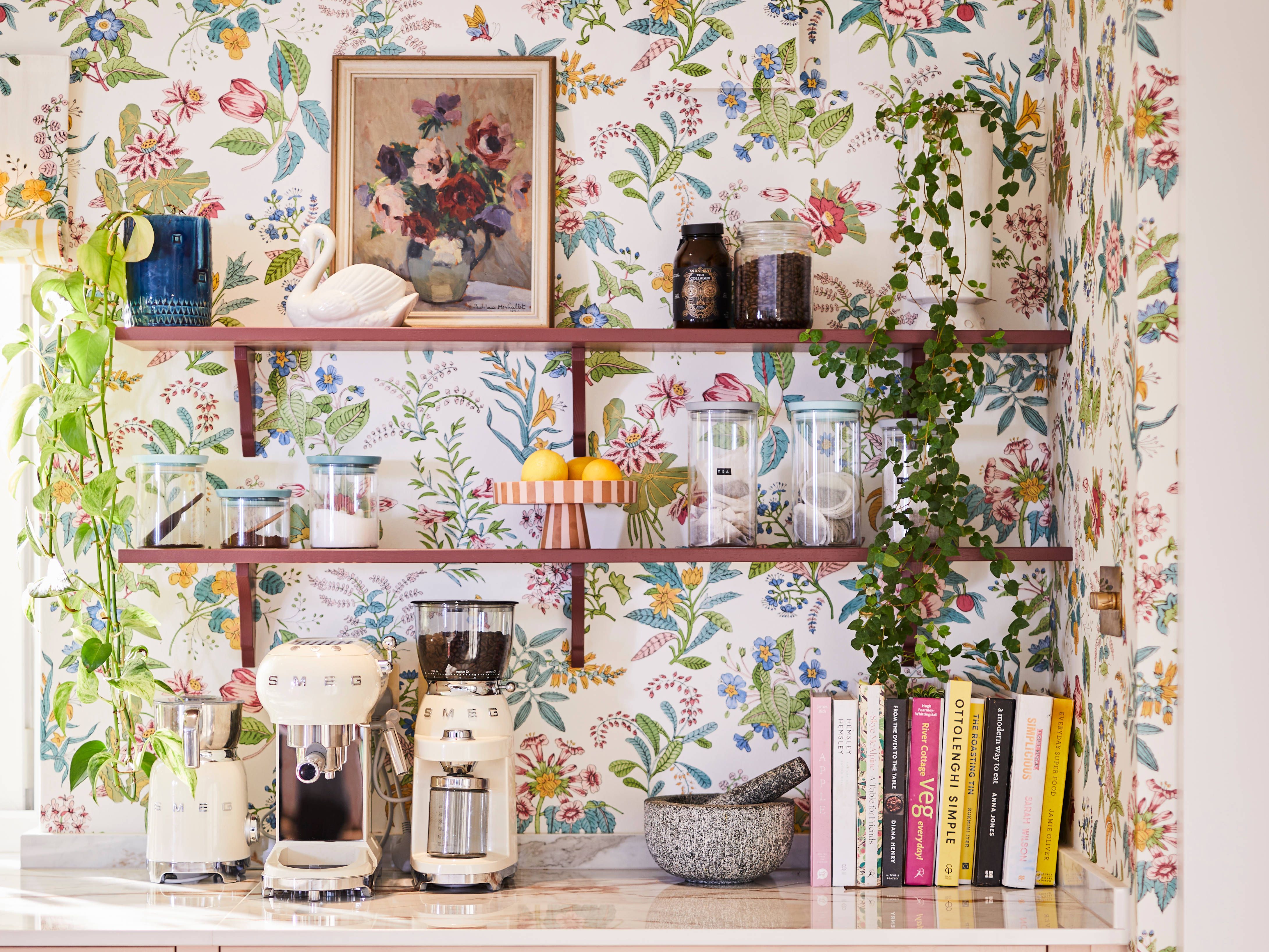 Coffee area in Sophie Robinson's maximalist kitchen, with shaker style Ludlow Chalk blush cabinetry, open shelving on a wall with a floral wallpaper and Smeg coffee machines on countertop.