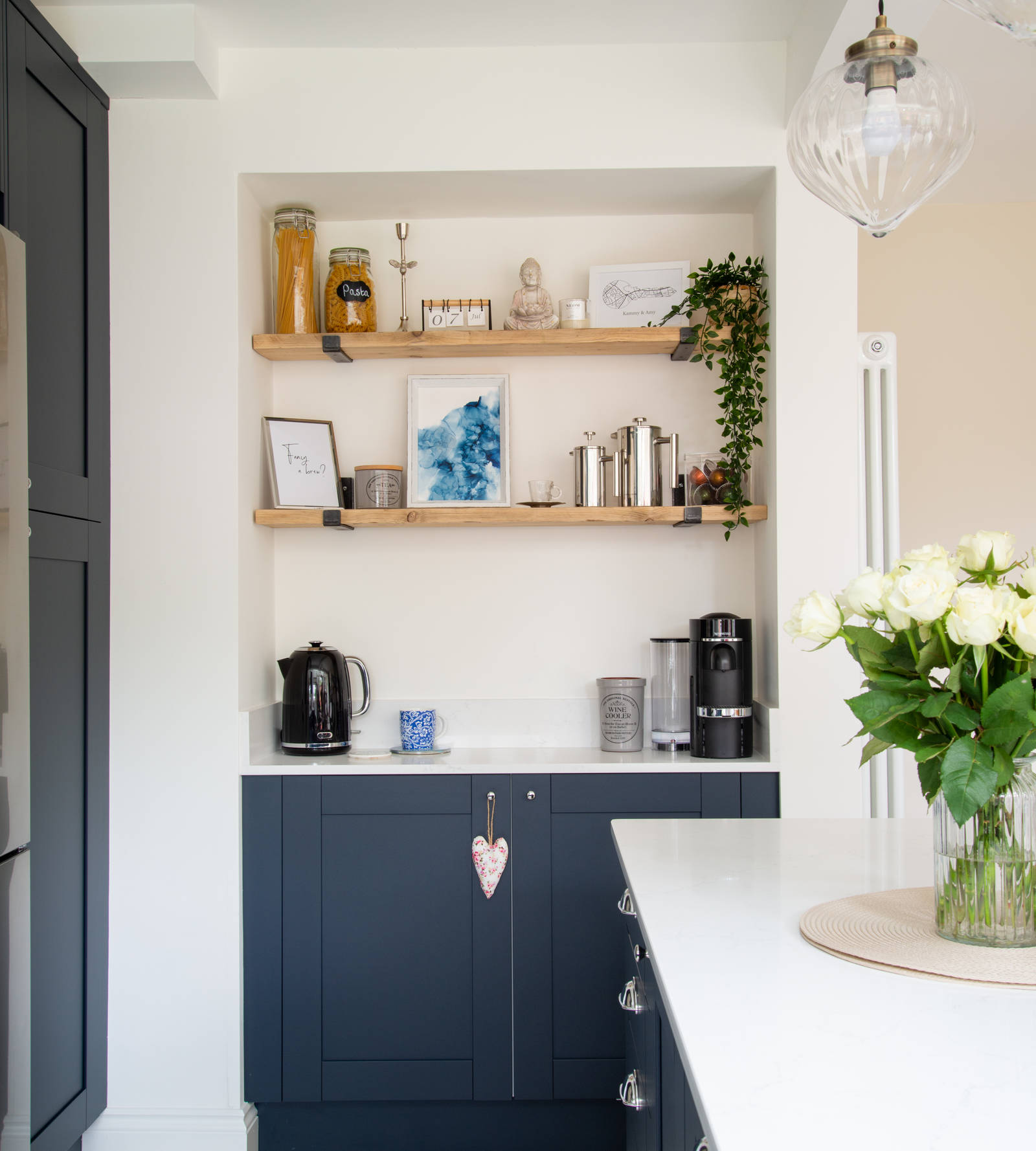 Dark blue shaker cabinets, white worktop, kettel and coffee machine with open wooden shelves on wall above.