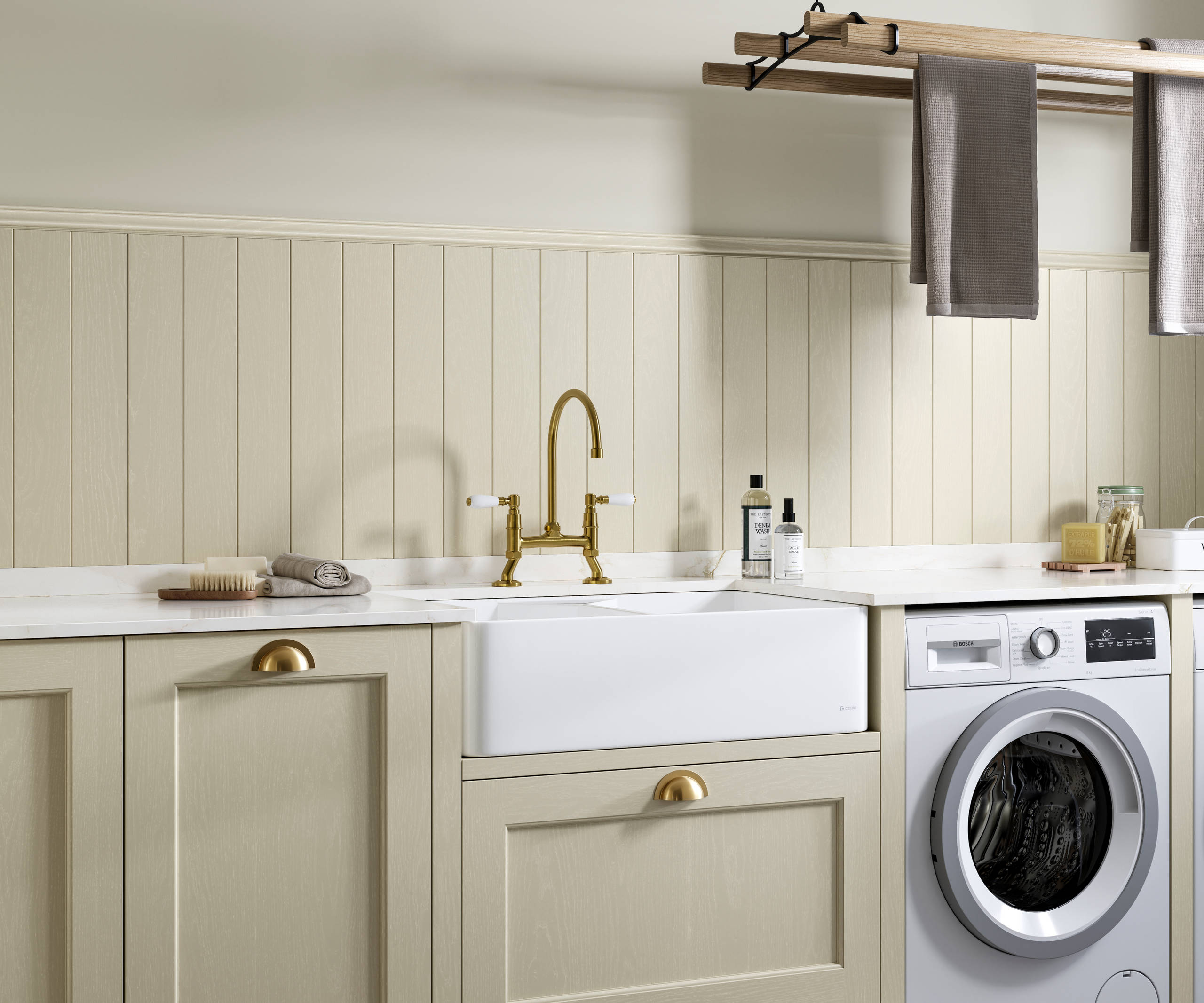 Traditional Shaker-style Ludlow Parchment and French Clay utility room with white ceramic sink, washing machine and tumble dryer with white marbled Silestone worktop and upstand, wallpanel matching the cabinetry, traditional brass tap and towels hanging on wood drying rack.