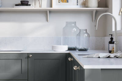 Ludlow, a traditional Shaker-style premium feel kitchen range with a visible woodgrain on its panelled doors here shown in the green shade Arboretum styled with steel knobs, steel tap, a white marbled worktop with matching splashback.