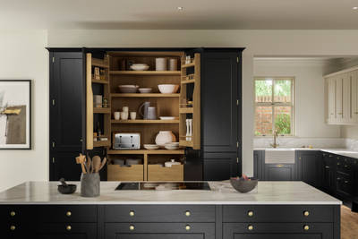 View over kitchen island in Traditional Shaker-style tactile woodgrain Ludlow Sumi Black kitchen, opened tall oak larder cabinets, brass accessories, marbled white and grey worktop, white ceramic sink and brass tap.